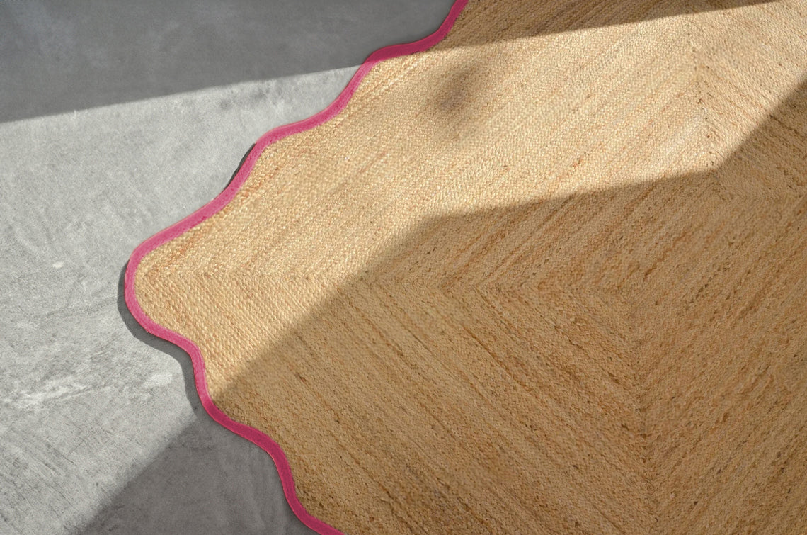 Handwoven natural jute scalloped area rug with pink border placed in a modern living room featuring a charcoal grey sofa.