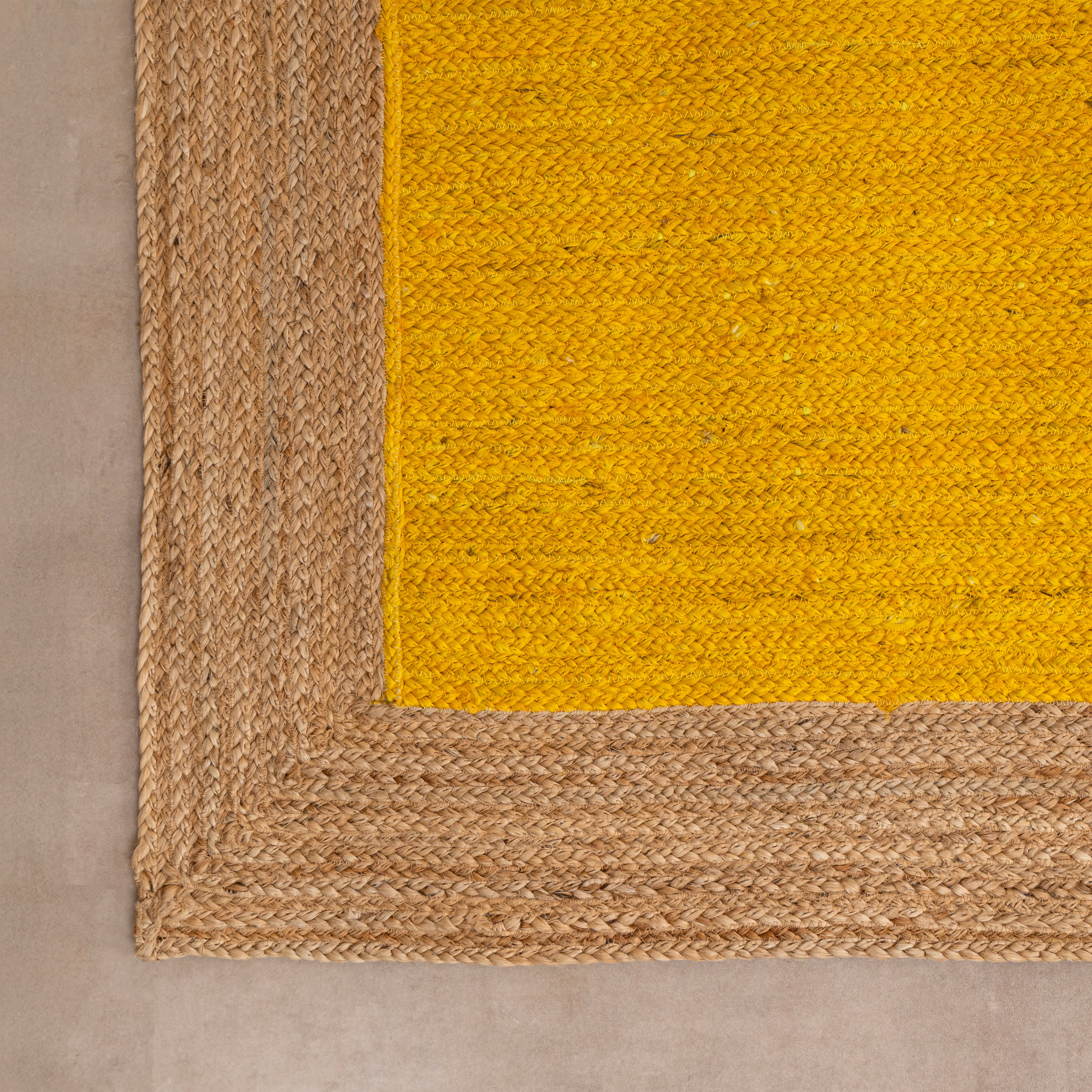 Handwoven mustard yellow natural jute area rug with a braided tan border, laid flat on a light beige tiled floor alongside a ceramic vase with dried bunny tails and a book.