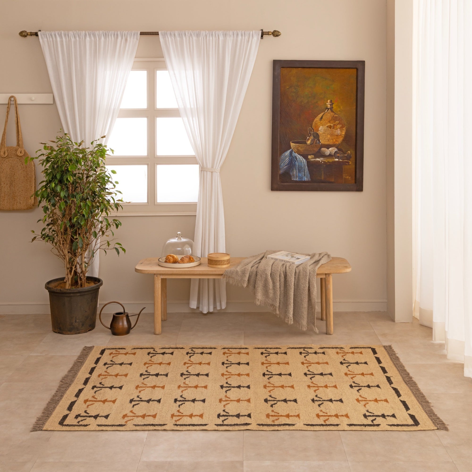 Handwoven natural jute kilim rug in beige with black and rust geometric tribal patterns, fringed edges, styled with a vase and book in a warm neutral living space.