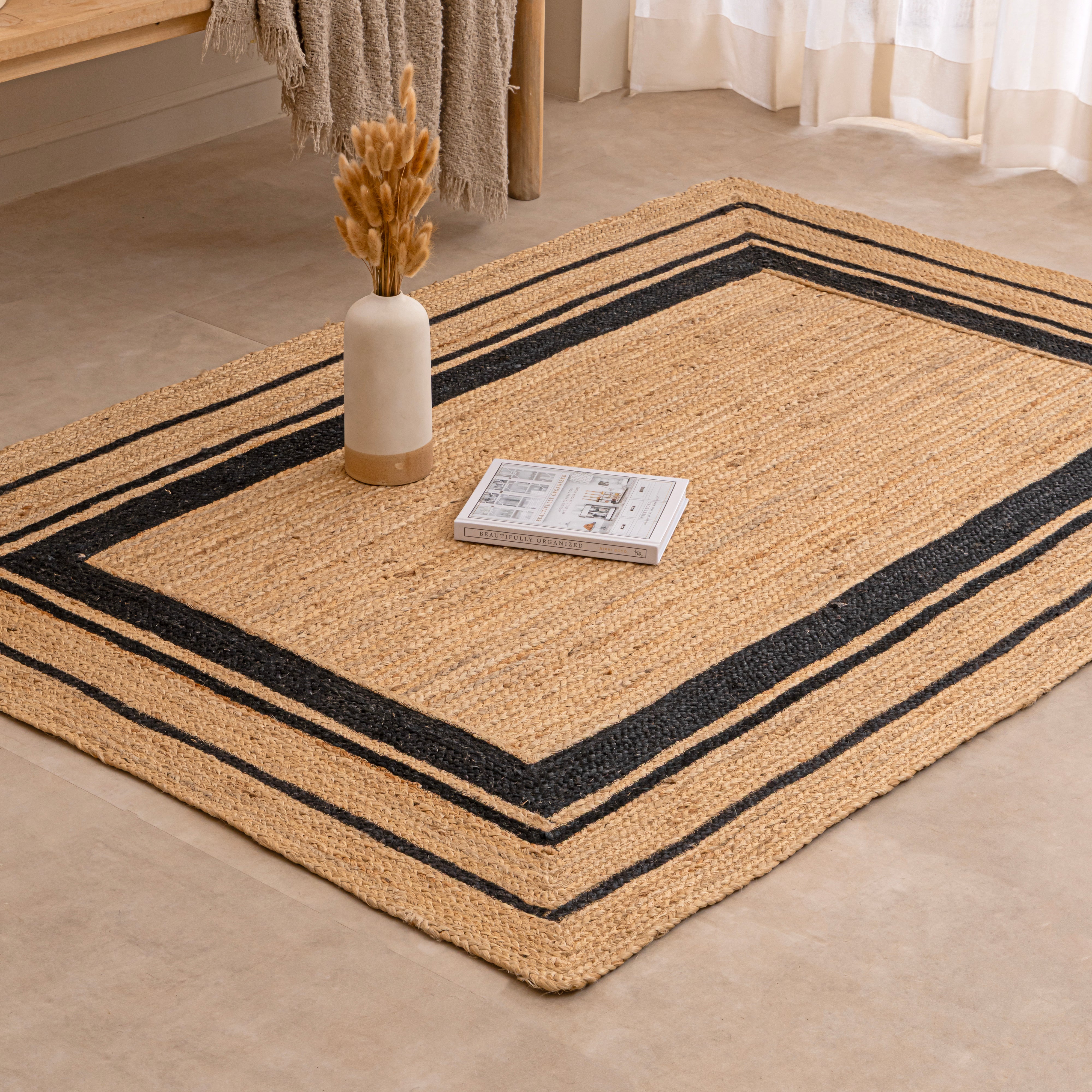 Handwoven natural tan jute area rug featuring a black double-striped border design, styled flat on a beige tiled floor next to a white ceramic vase with dried bunny tails and a hardcover book.
