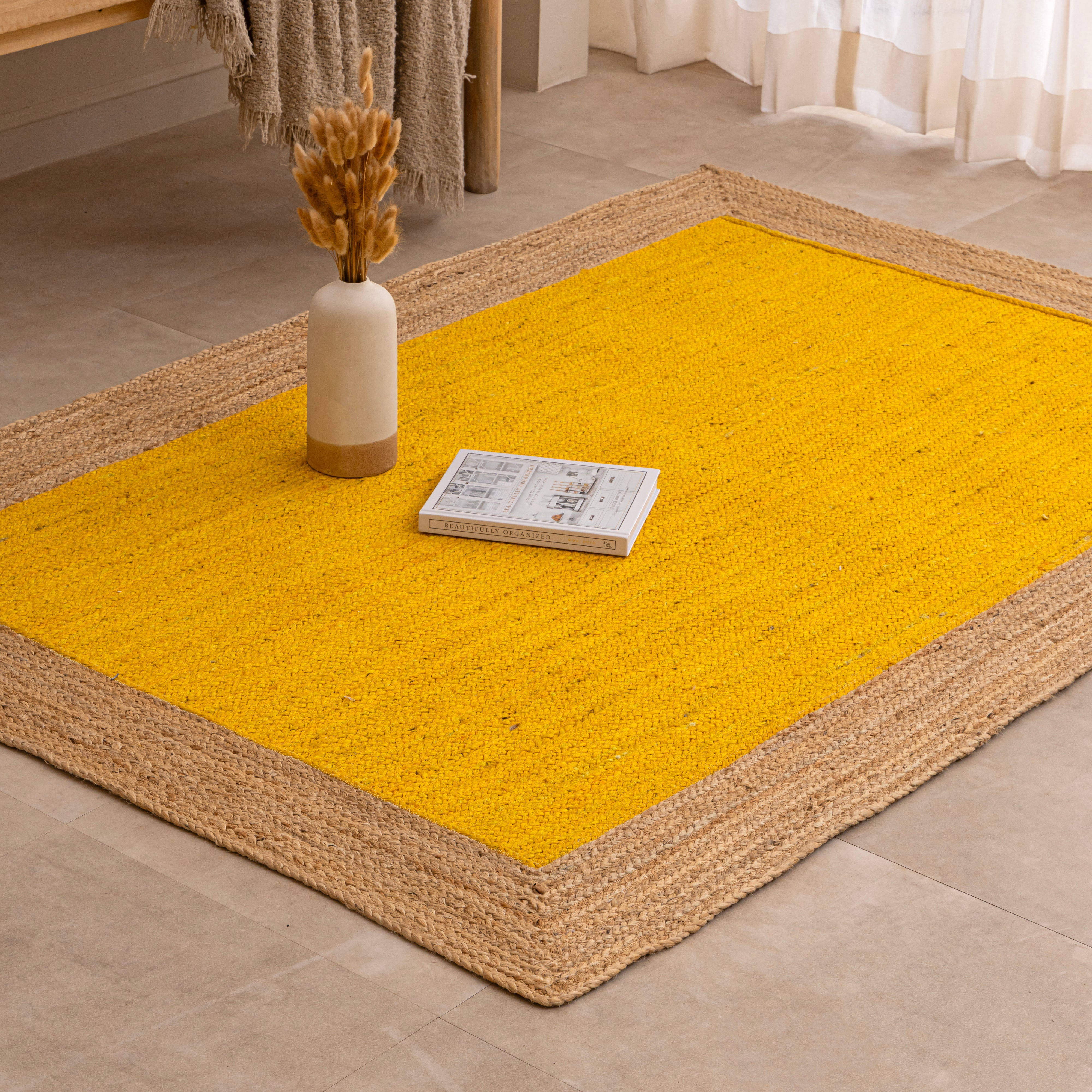 Handwoven mustard yellow natural jute area rug with a braided tan border, laid flat on a light beige tiled floor alongside a ceramic vase with dried bunny tails and a book.