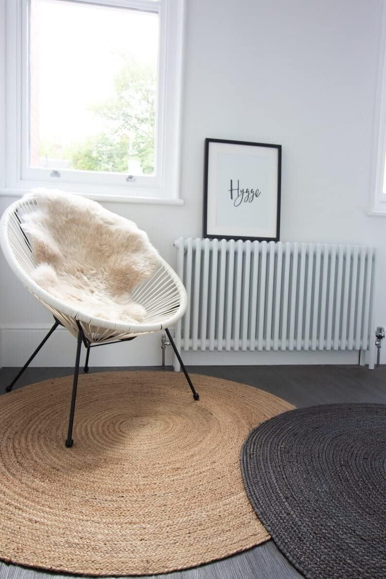 A handwoven, round natural jute area rug placed on a light wood floor in a cozy, brightly lit modern living room with a beige sofa and woven decor.