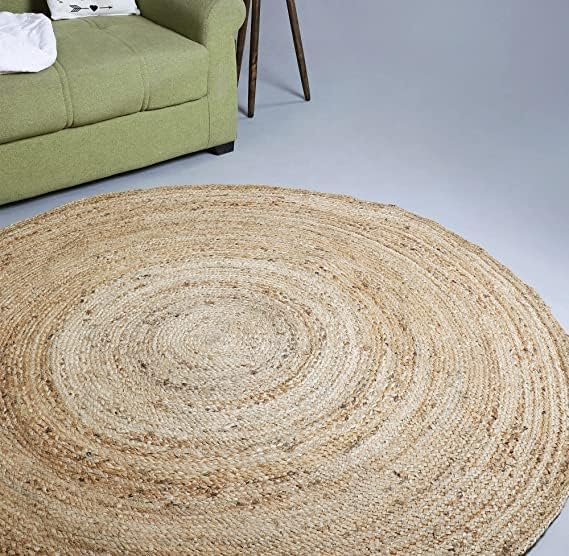 A handwoven, round natural jute area rug placed on a light wood floor in a cozy, brightly lit modern living room with a beige sofa and woven decor.
