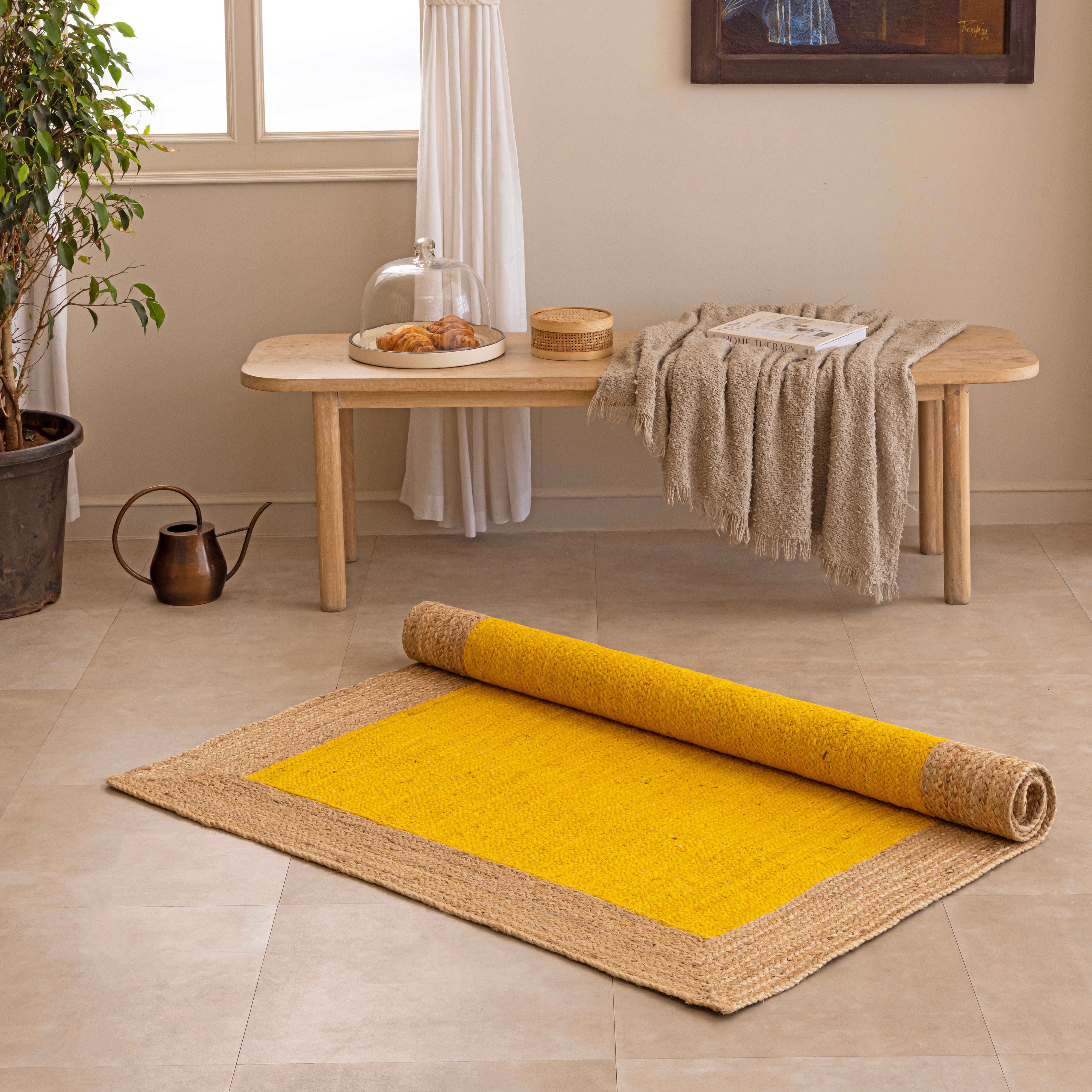 Handwoven mustard yellow natural jute area rug with a braided tan border, laid flat on a light beige tiled floor alongside a ceramic vase with dried bunny tails and a book.