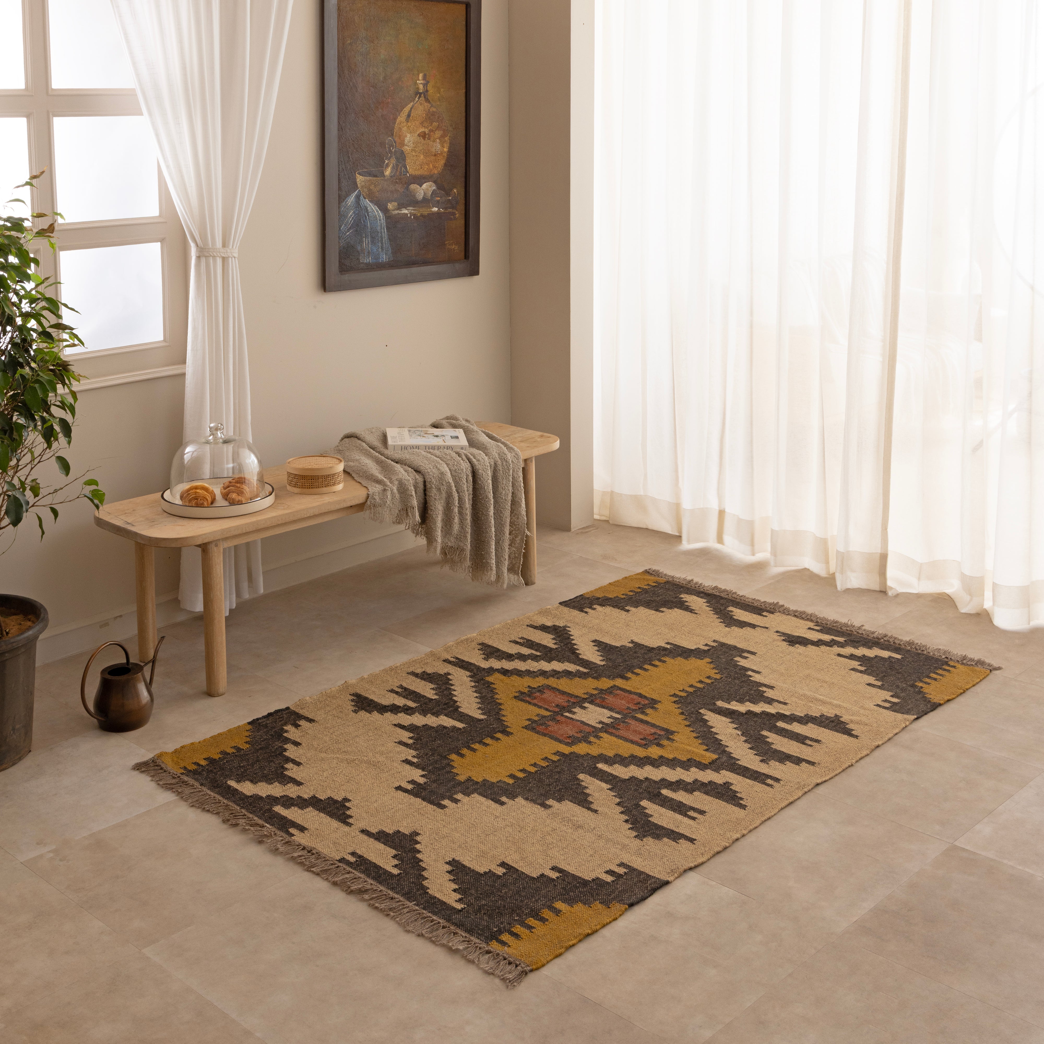 Handwoven Southwestern style natural jute kilim rug with geometric black and yellow patterns and fringed edges, styled on a beige tiled floor in front of a wooden bench and large indoor plant.