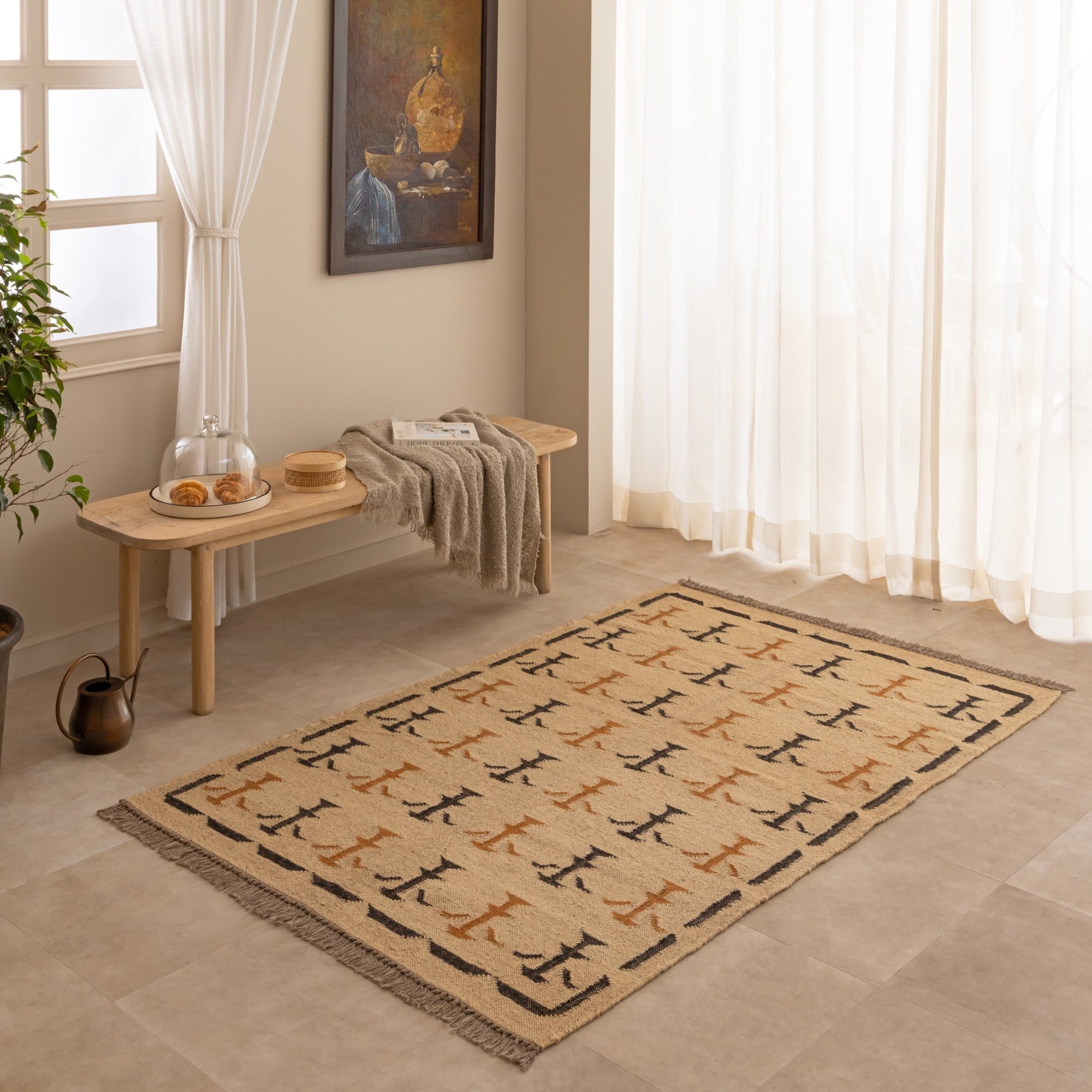 Handwoven natural jute kilim rug in beige with black and rust geometric tribal patterns, fringed edges, styled with a vase and book in a warm neutral living space.
