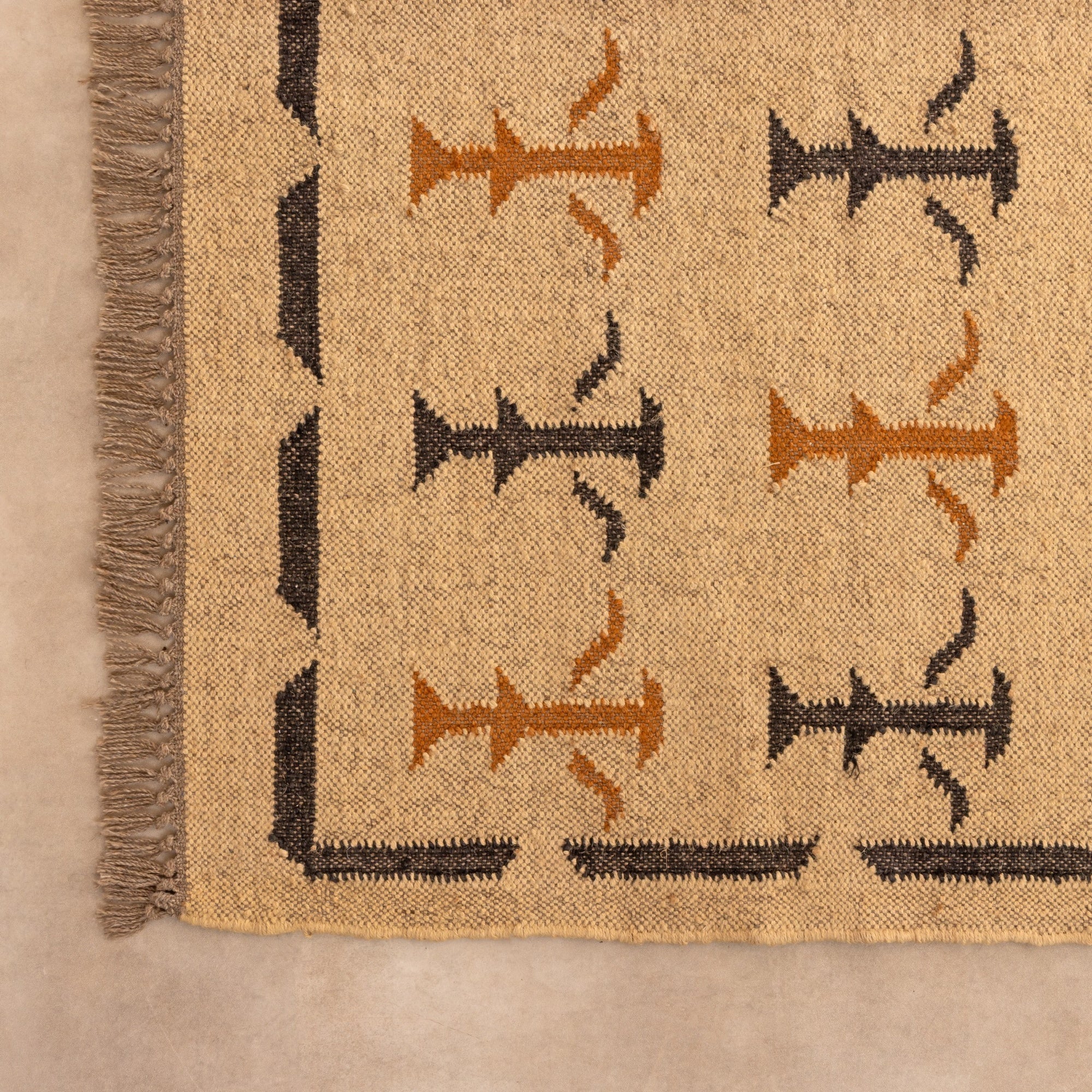 Handwoven natural jute kilim rug in beige with black and rust geometric tribal patterns, fringed edges, styled with a vase and book in a warm neutral living space.