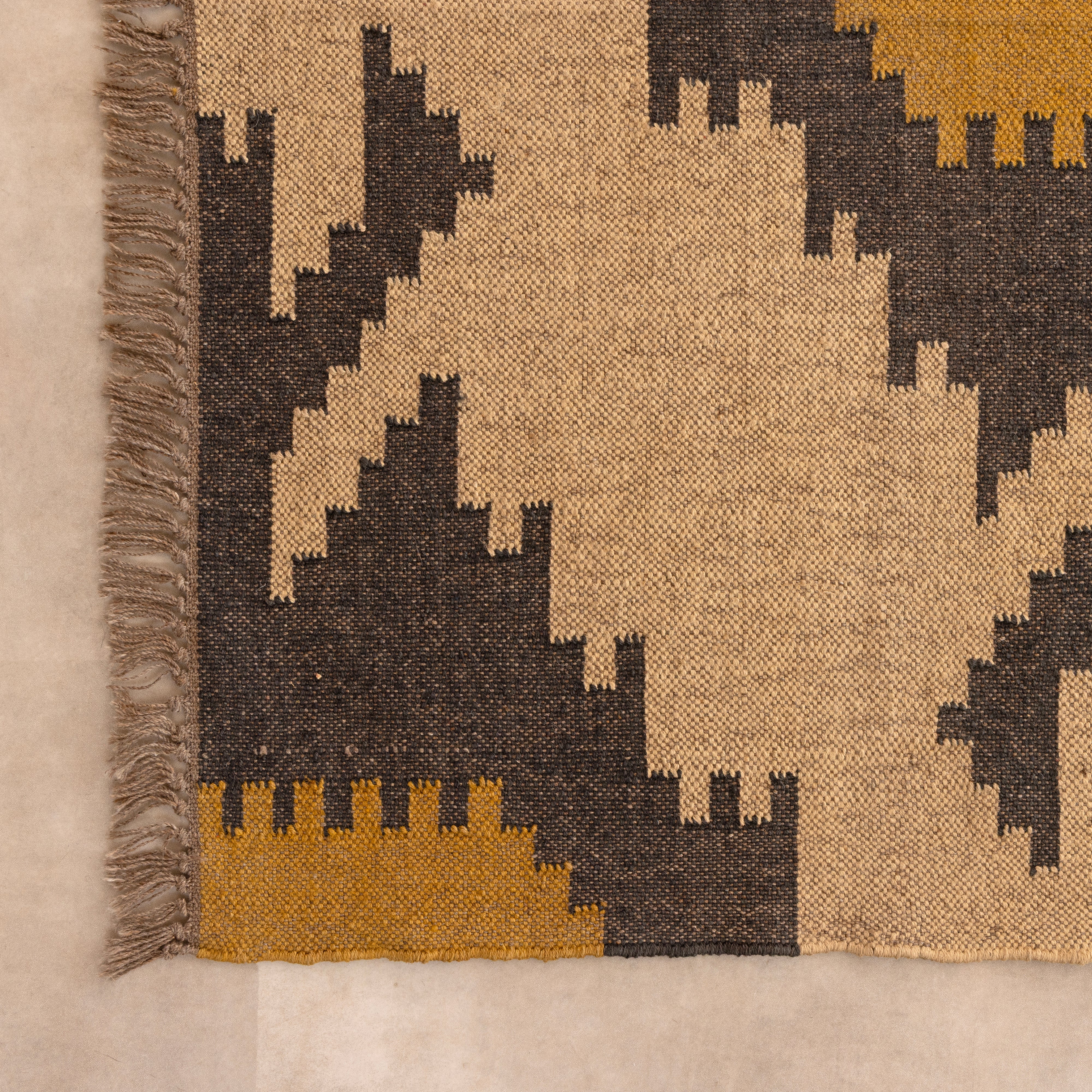 Handwoven Southwestern style natural jute kilim rug with geometric black and yellow patterns and fringed edges, styled on a beige tiled floor in front of a wooden bench and large indoor plant.
