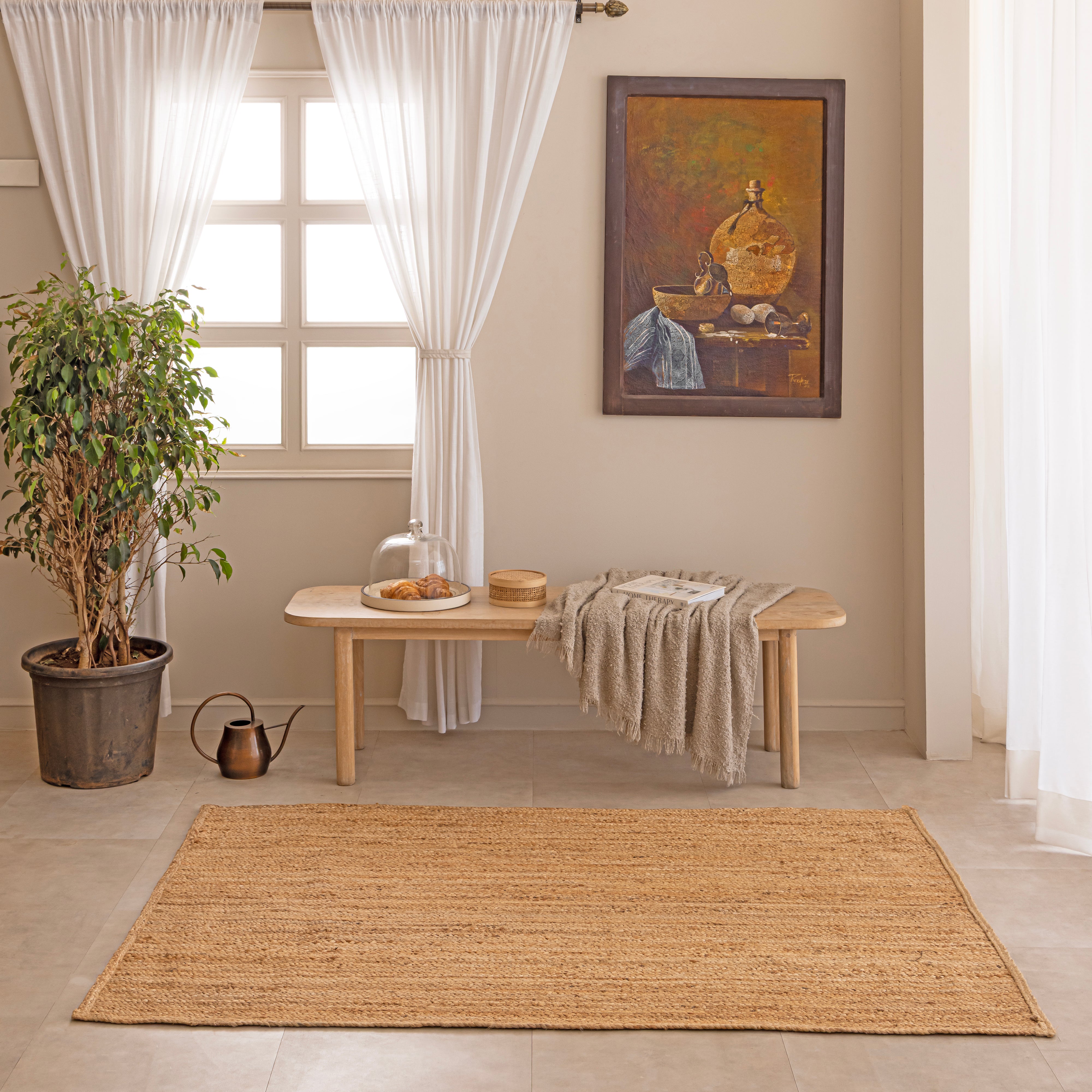Handcrafted solid natural tan jute area rug featuring a thick braided texture, laid flat on a beige tiled floor beside a minimalist white ceramic vase with dried bunny tails and a hardcover book.