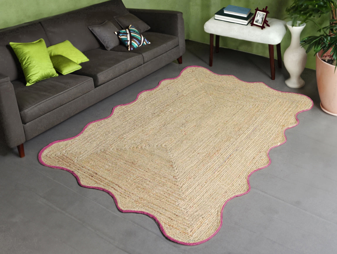 Handwoven natural jute scalloped area rug with pink border placed in a modern living room featuring a charcoal grey sofa.