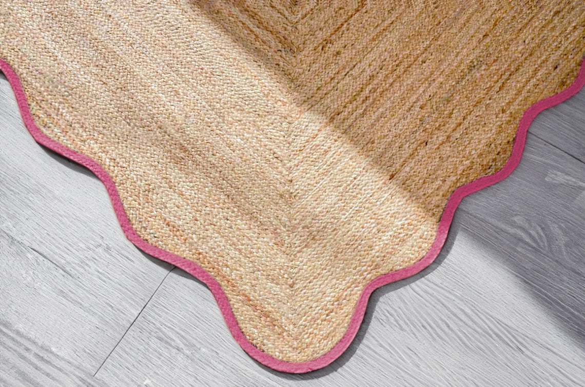 Handwoven natural jute scalloped area rug with pink border placed in a modern living room featuring a charcoal grey sofa.