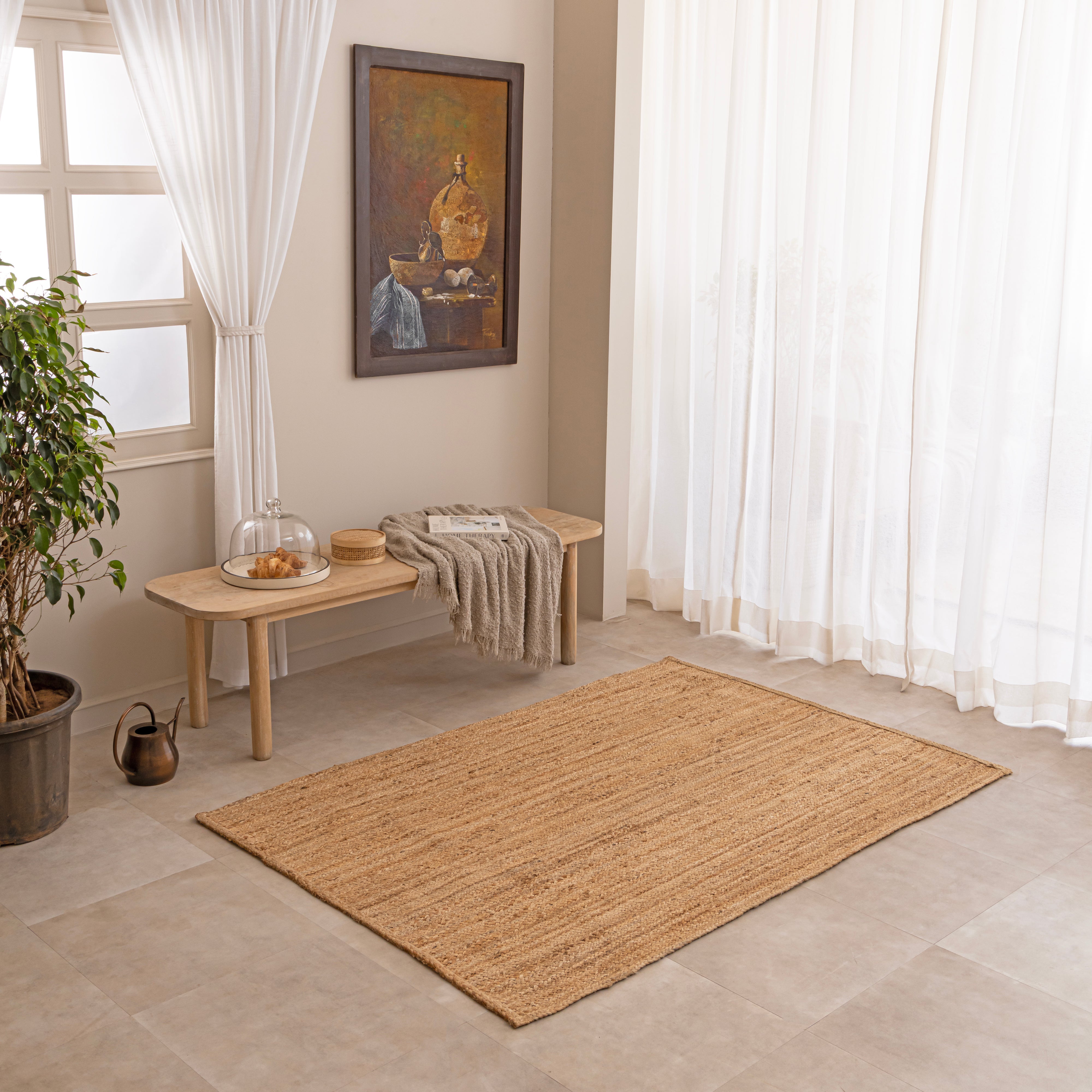 Handcrafted solid natural tan jute area rug featuring a thick braided texture, laid flat on a beige tiled floor beside a minimalist white ceramic vase with dried bunny tails and a hardcover book.