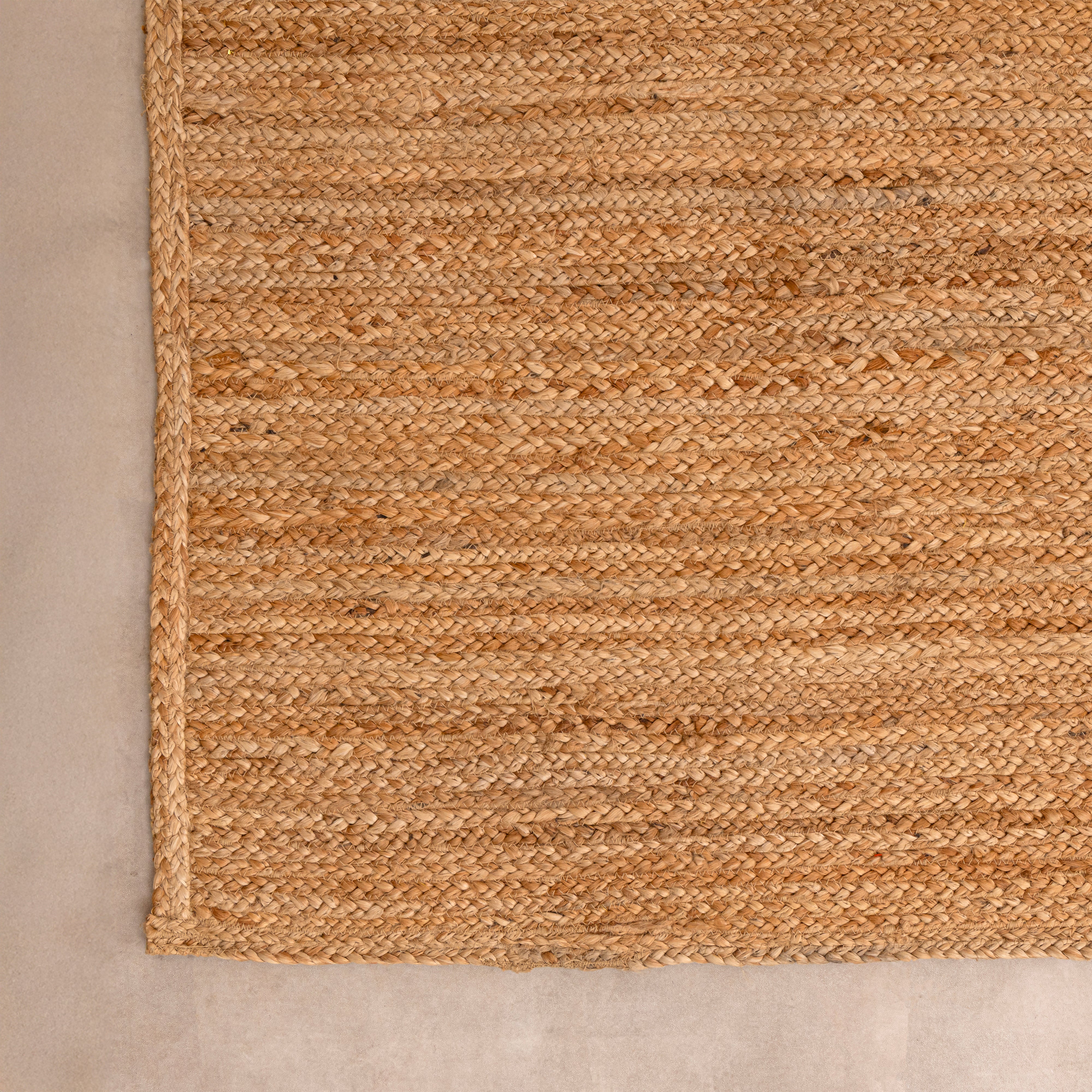 Handcrafted solid natural tan jute area rug featuring a thick braided texture, laid flat on a beige tiled floor beside a minimalist white ceramic vase with dried bunny tails and a hardcover book.