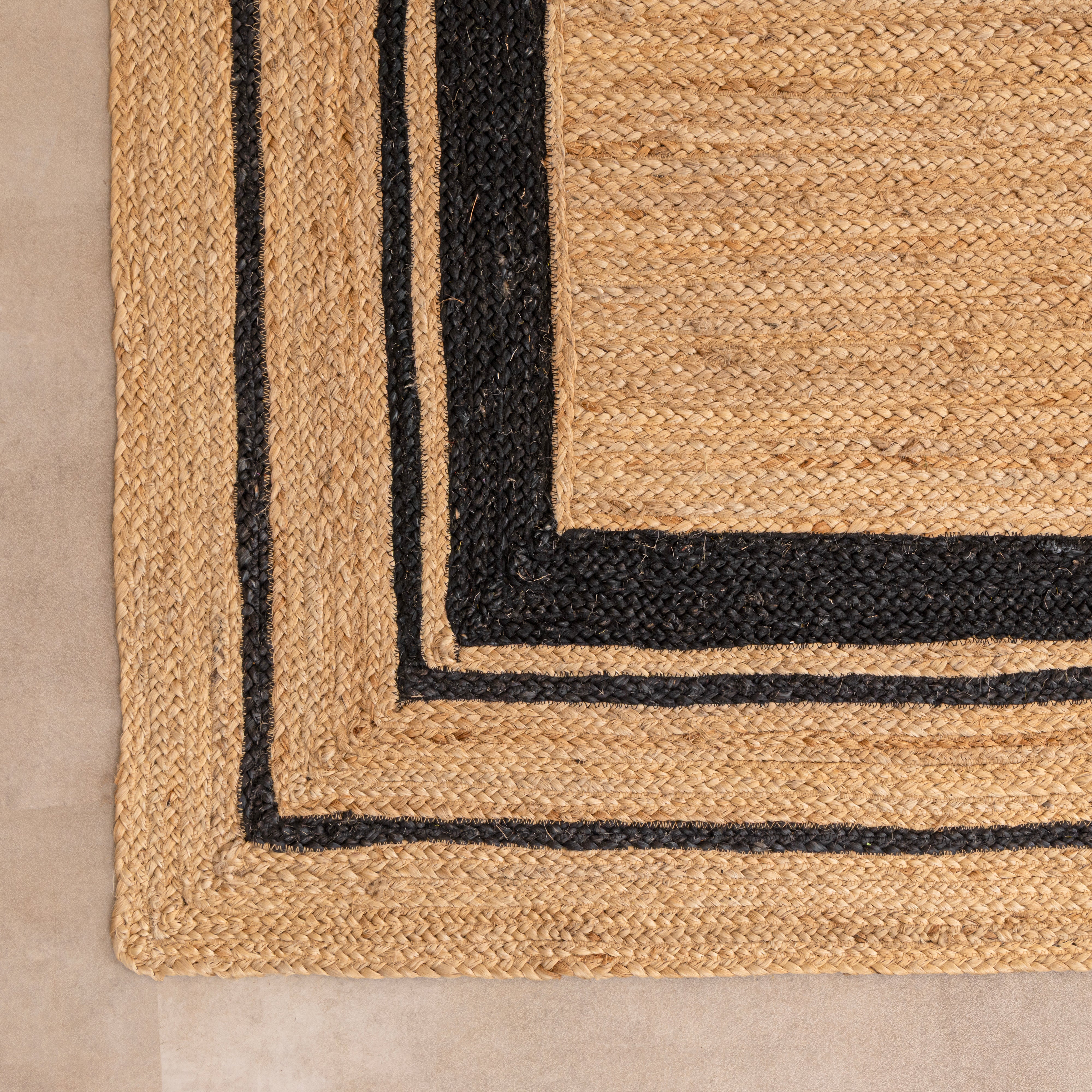 Handwoven natural tan jute area rug featuring a black double-striped border design, styled flat on a beige tiled floor next to a white ceramic vase with dried bunny tails and a hardcover book.