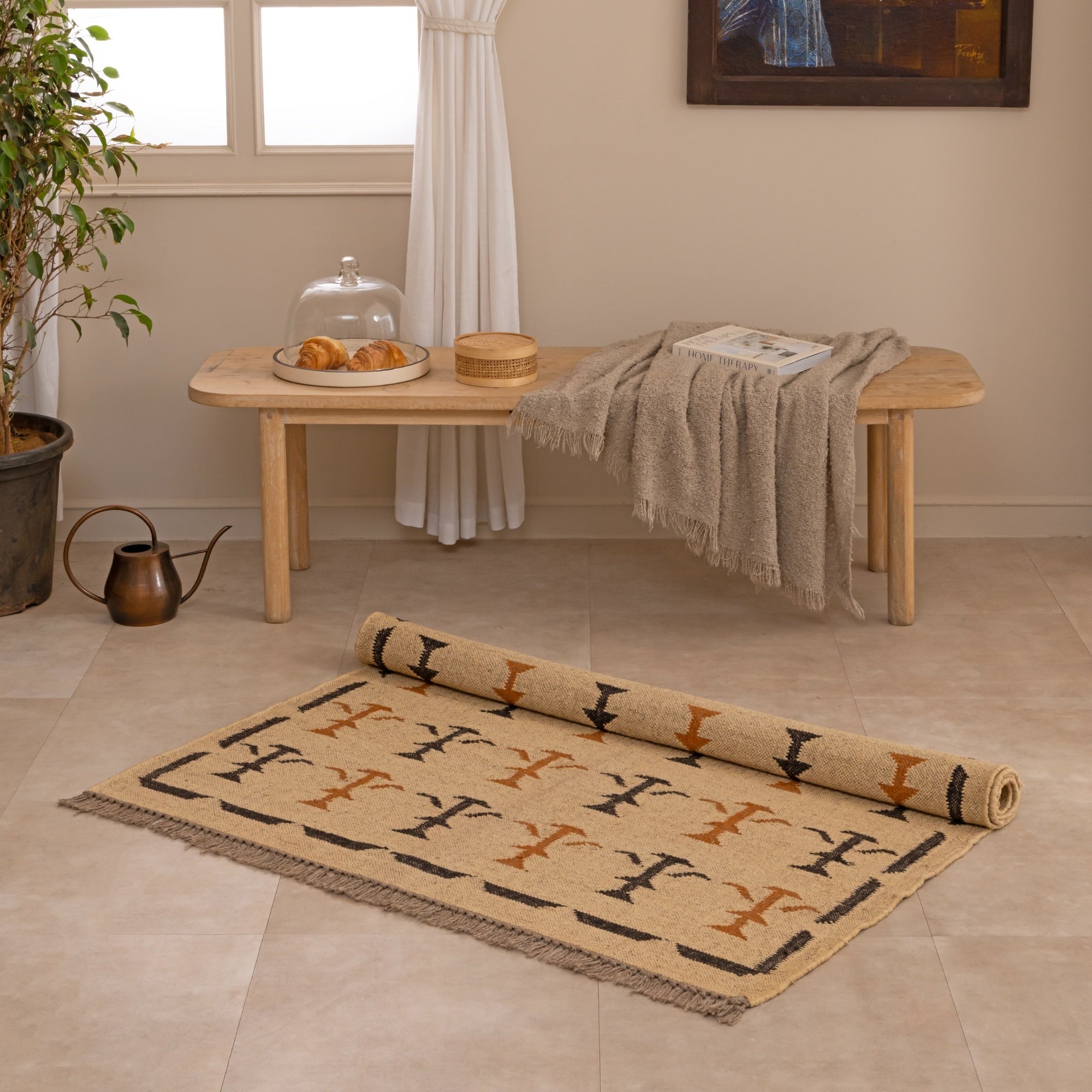 Handwoven natural jute kilim rug in beige with black and rust geometric tribal patterns, fringed edges, styled with a vase and book in a warm neutral living space.
