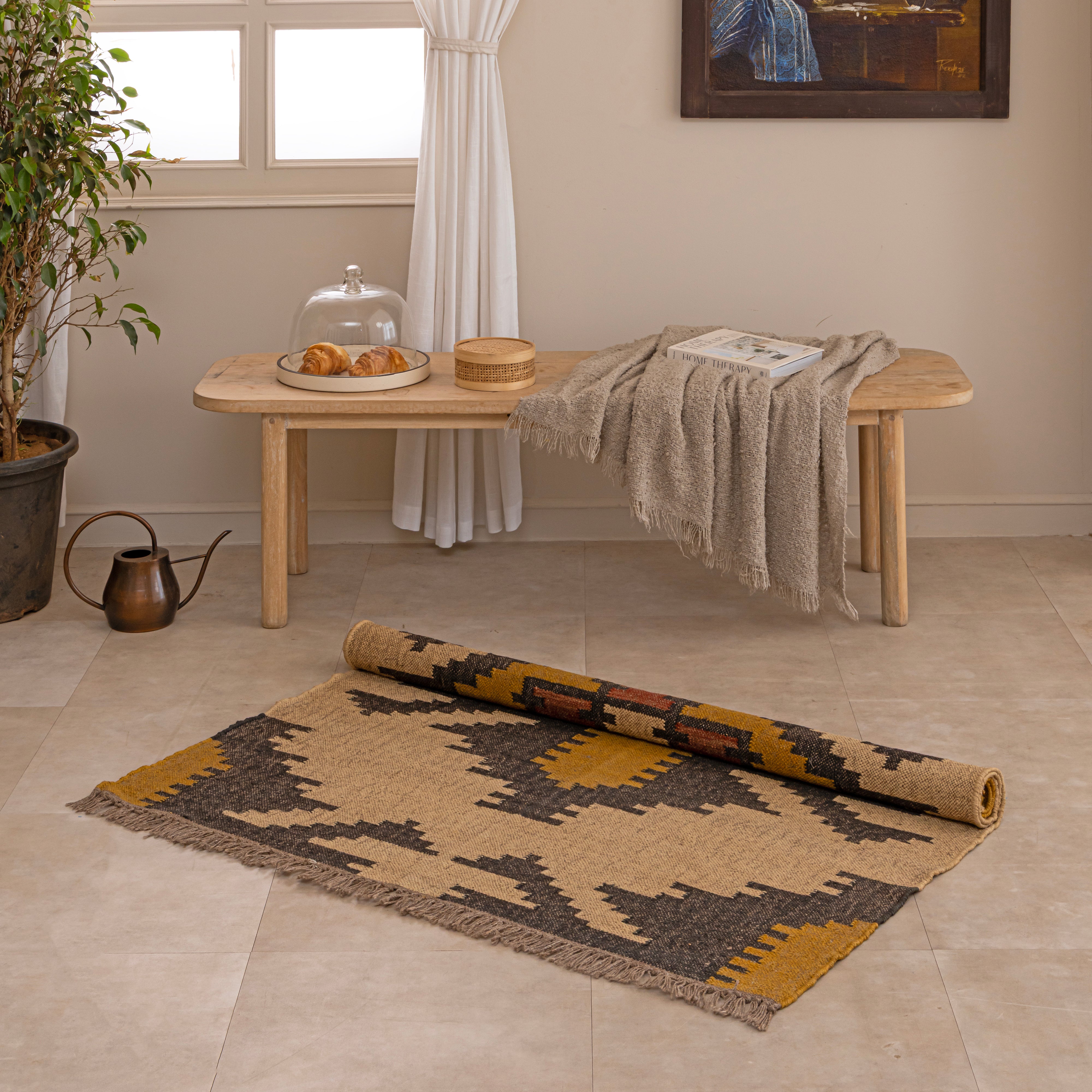 Handwoven Southwestern style natural jute kilim rug with geometric black and yellow patterns and fringed edges, styled on a beige tiled floor in front of a wooden bench and large indoor plant.