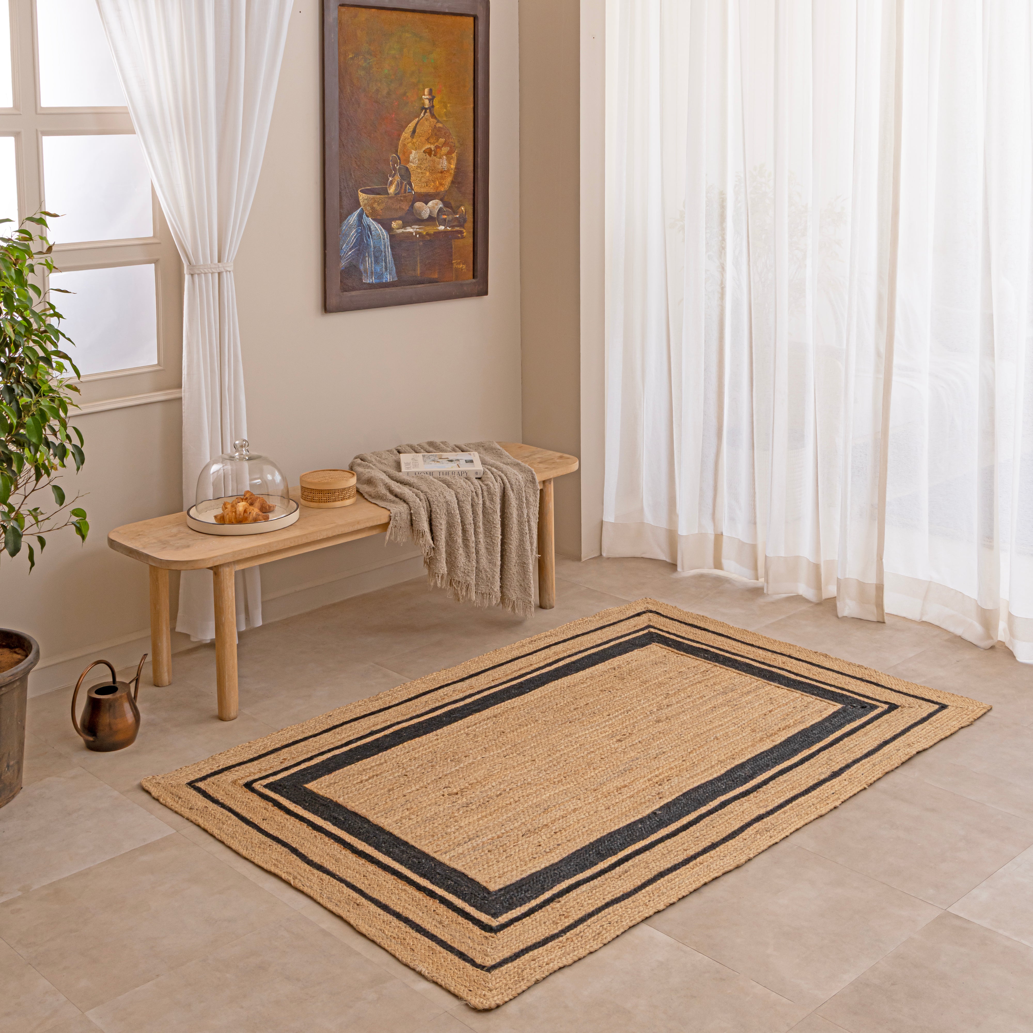 Handwoven natural tan jute area rug featuring a black double-striped border design, styled flat on a beige tiled floor next to a white ceramic vase with dried bunny tails and a hardcover book.