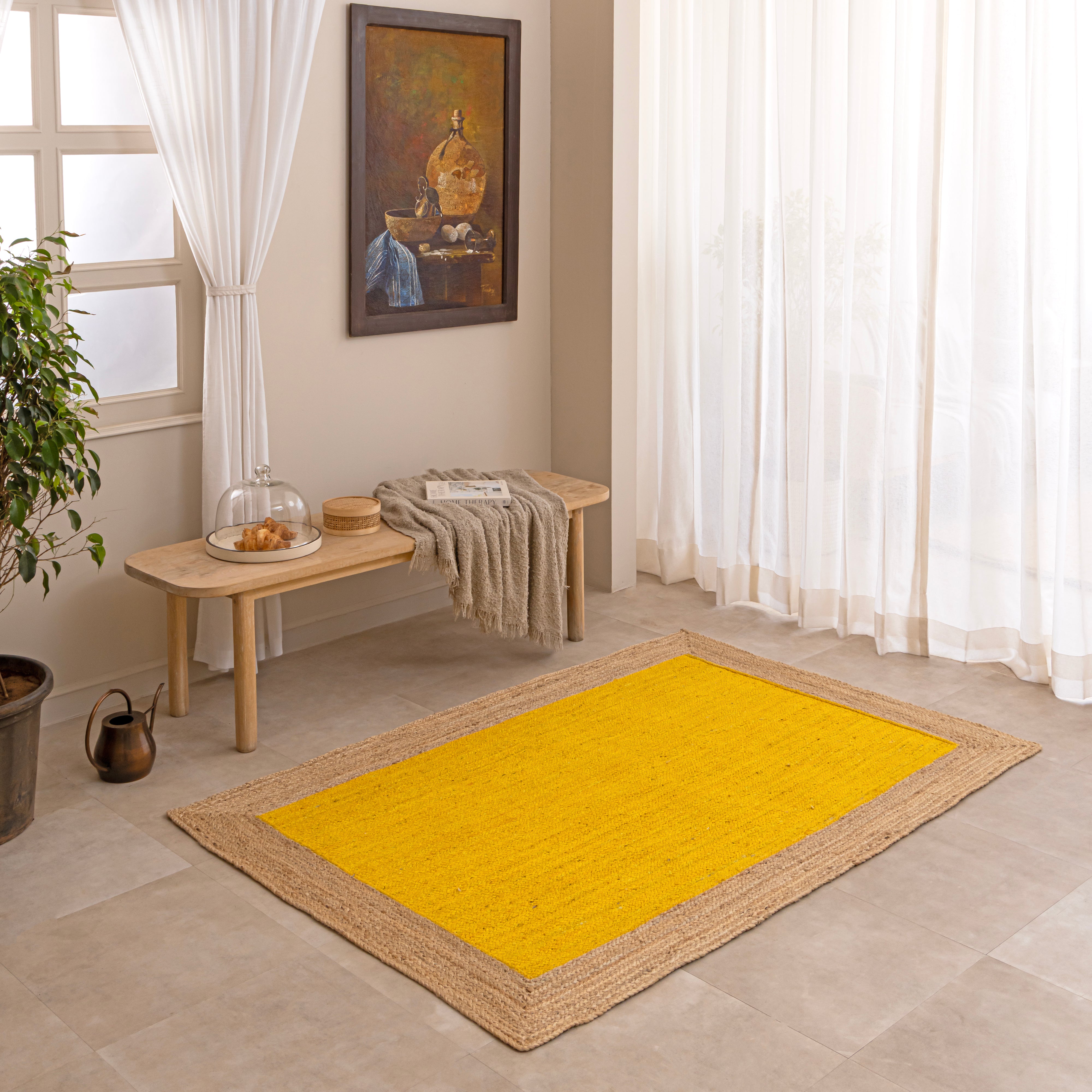 Handwoven mustard yellow natural jute area rug with a braided tan border, laid flat on a light beige tiled floor alongside a ceramic vase with dried bunny tails and a book.