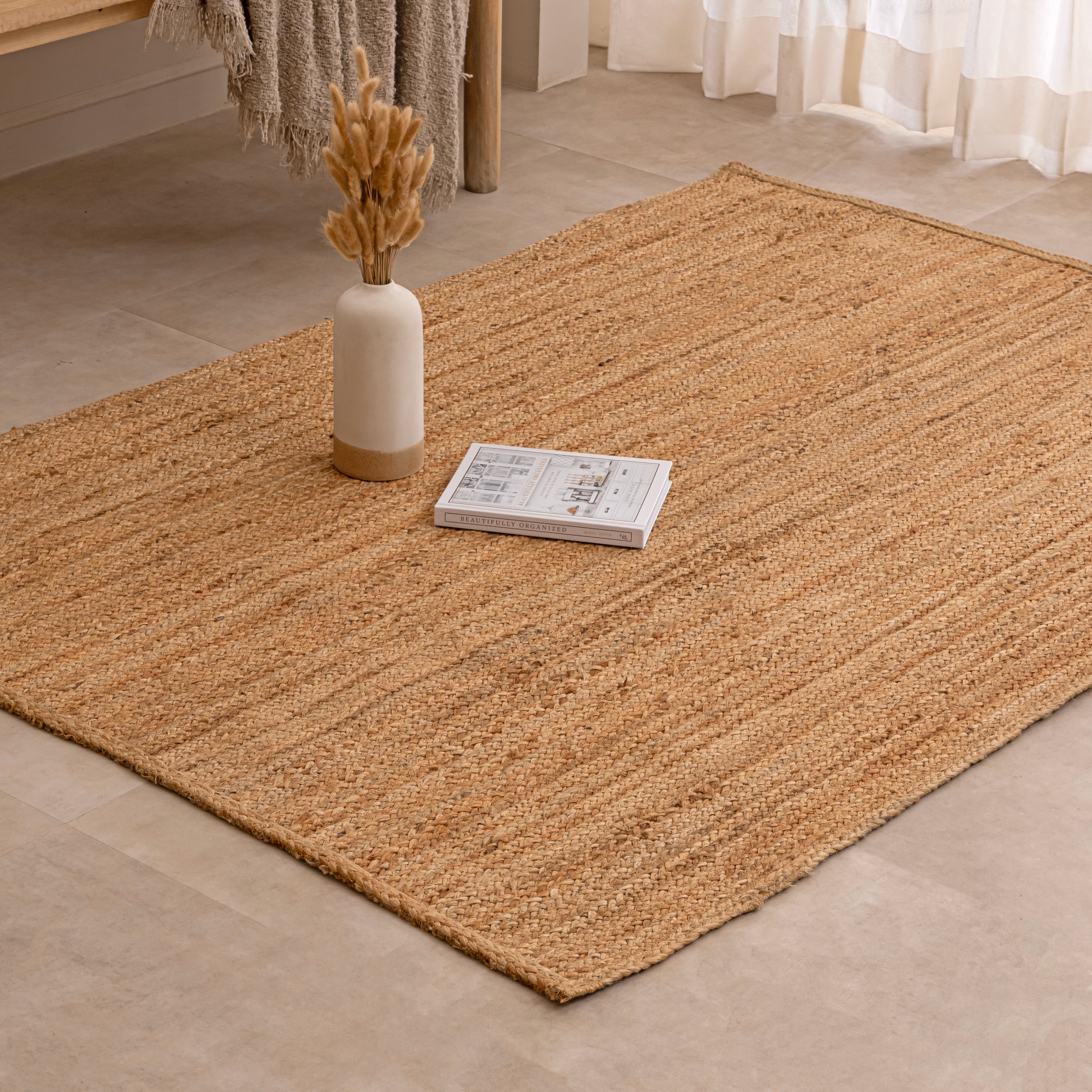 Handcrafted solid natural tan jute area rug featuring a thick braided texture, laid flat on a beige tiled floor beside a minimalist white ceramic vase with dried bunny tails and a hardcover book.