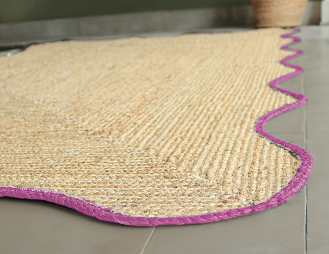 Handwoven natural jute scalloped area rug with pink border placed in a modern living room featuring a charcoal grey sofa.