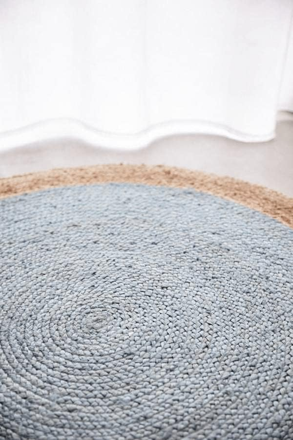 A two-tone round jute area rug featuring a muted light blue braided center and a natural tan woven border, laid on a light wood floor in a cozy coastal living room.