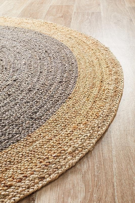 A charcoal grey and natural tan two-tone round braided jute rug on a wooden floor in a warm living room with a beige sofa, sage green pillow, and potted plants