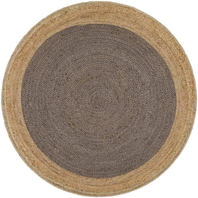 A charcoal grey and natural tan two-tone round braided jute rug on a wooden floor in a warm living room with a beige sofa, sage green pillow, and potted plants