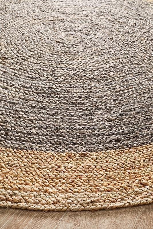 A charcoal grey and natural tan two-tone round braided jute rug on a wooden floor in a warm living room with a beige sofa, sage green pillow, and potted plants