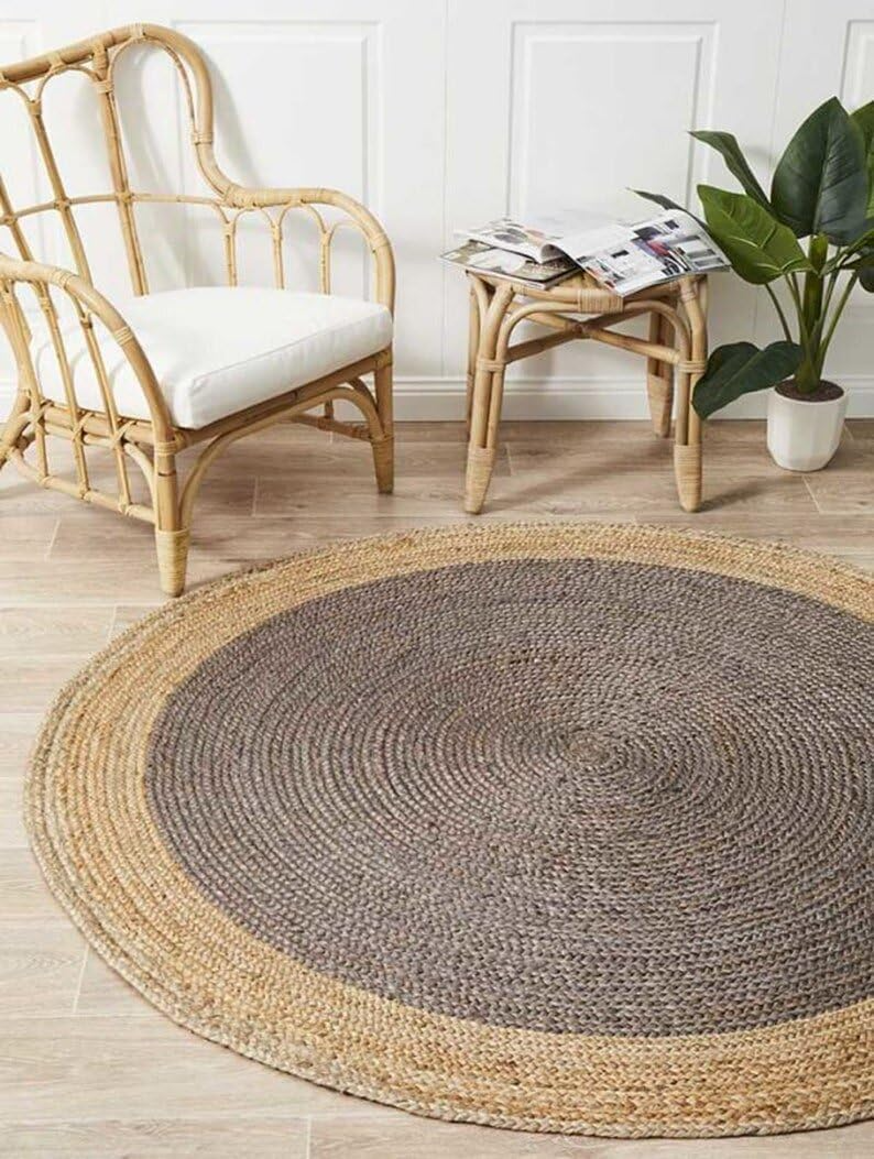 A charcoal grey and natural tan two-tone round braided jute rug on a wooden floor in a warm living room with a beige sofa, sage green pillow, and potted plants