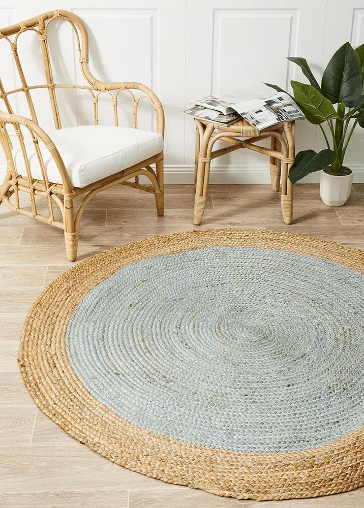 A two-tone round jute area rug featuring a muted light blue braided center and a natural tan woven border, laid on a light wood floor in a cozy coastal living room.