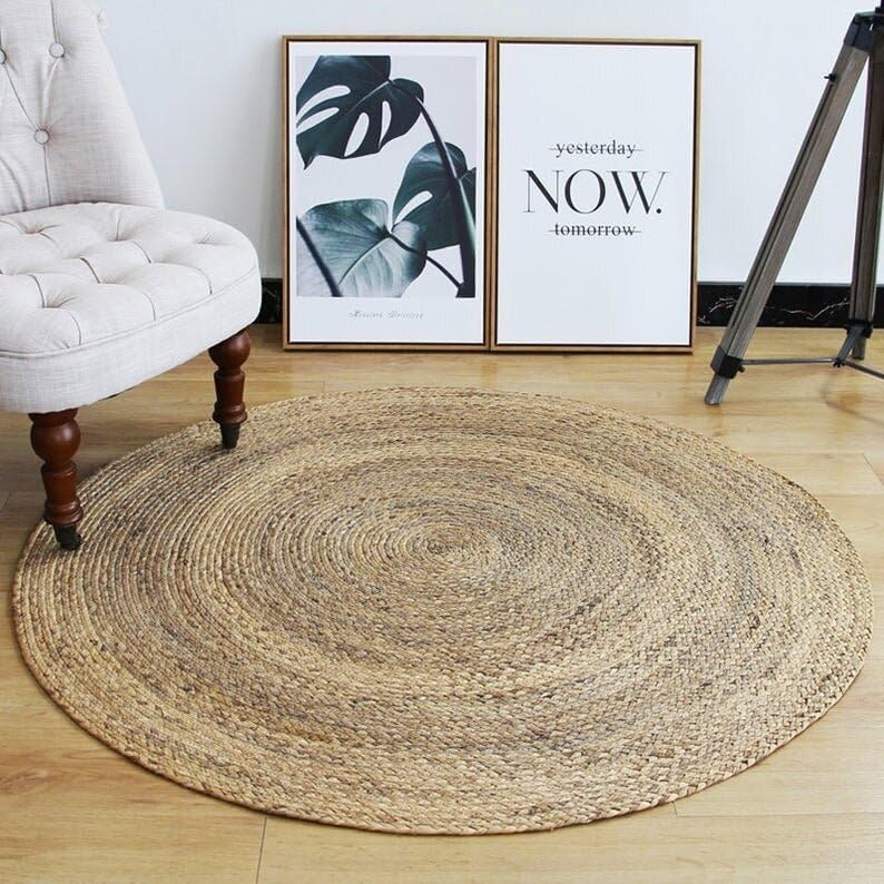 A handwoven, round natural jute area rug placed on a light wood floor in a cozy, brightly lit modern living room with a beige sofa and woven decor.