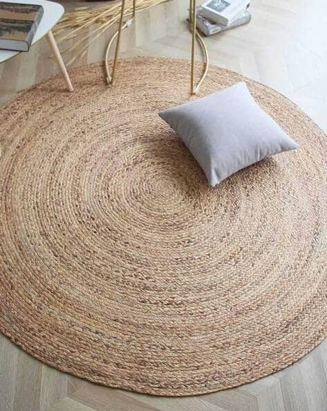 A handwoven, round natural jute area rug placed on a light wood floor in a cozy, brightly lit modern living room with a beige sofa and woven decor.