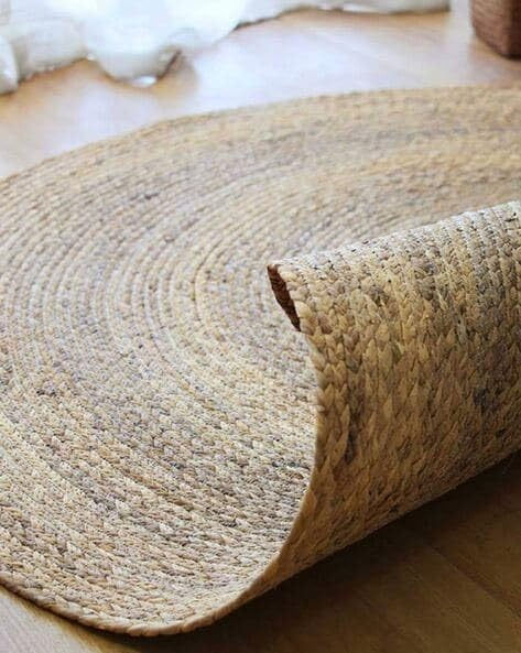 A handwoven, round natural jute area rug placed on a light wood floor in a cozy, brightly lit modern living room with a beige sofa and woven decor.