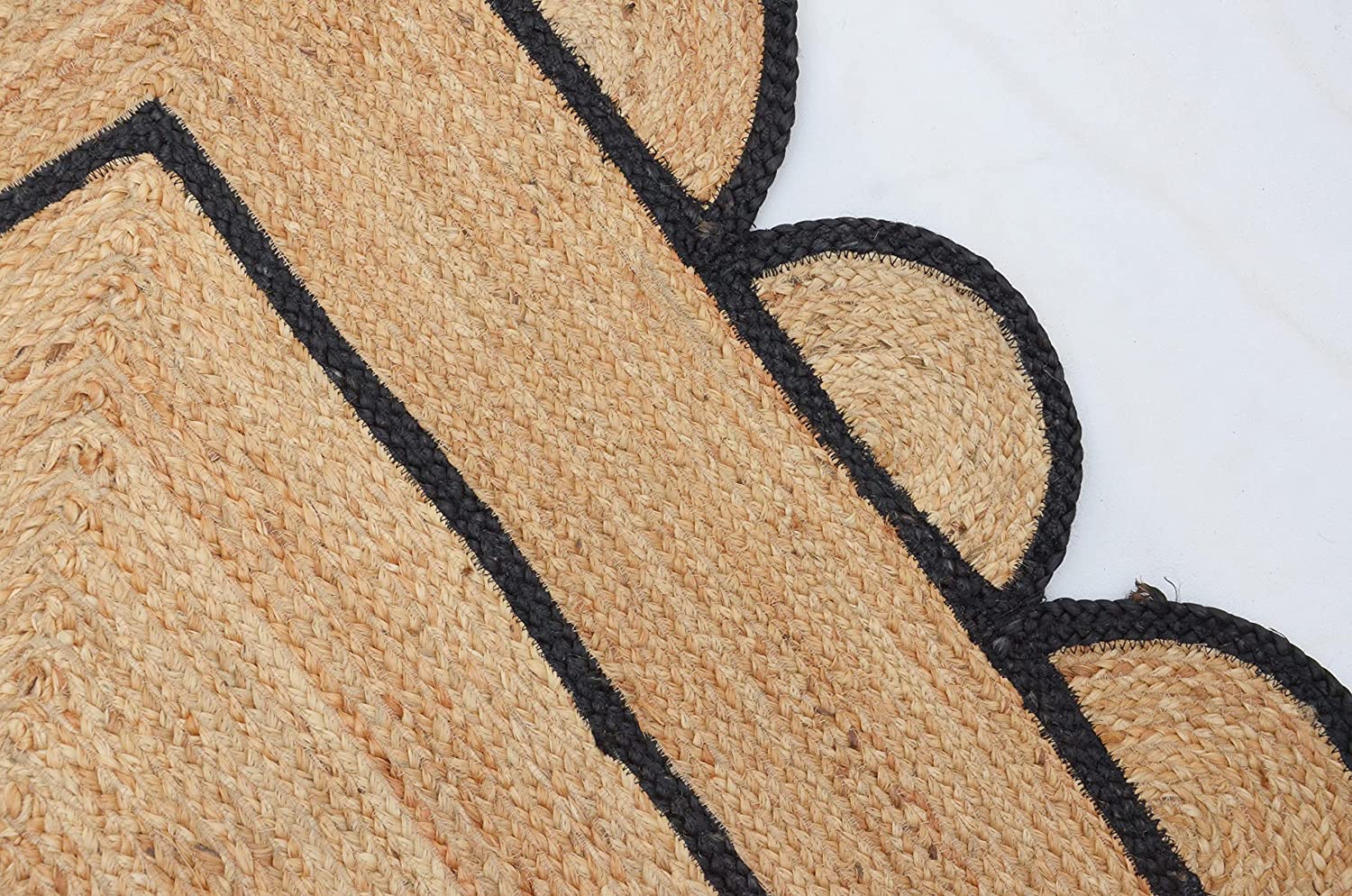 Handwoven natural jute area rug with black scalloped border in a modern Scandinavian living room featuring a cream sofa, wooden chair, and light wood flooring.