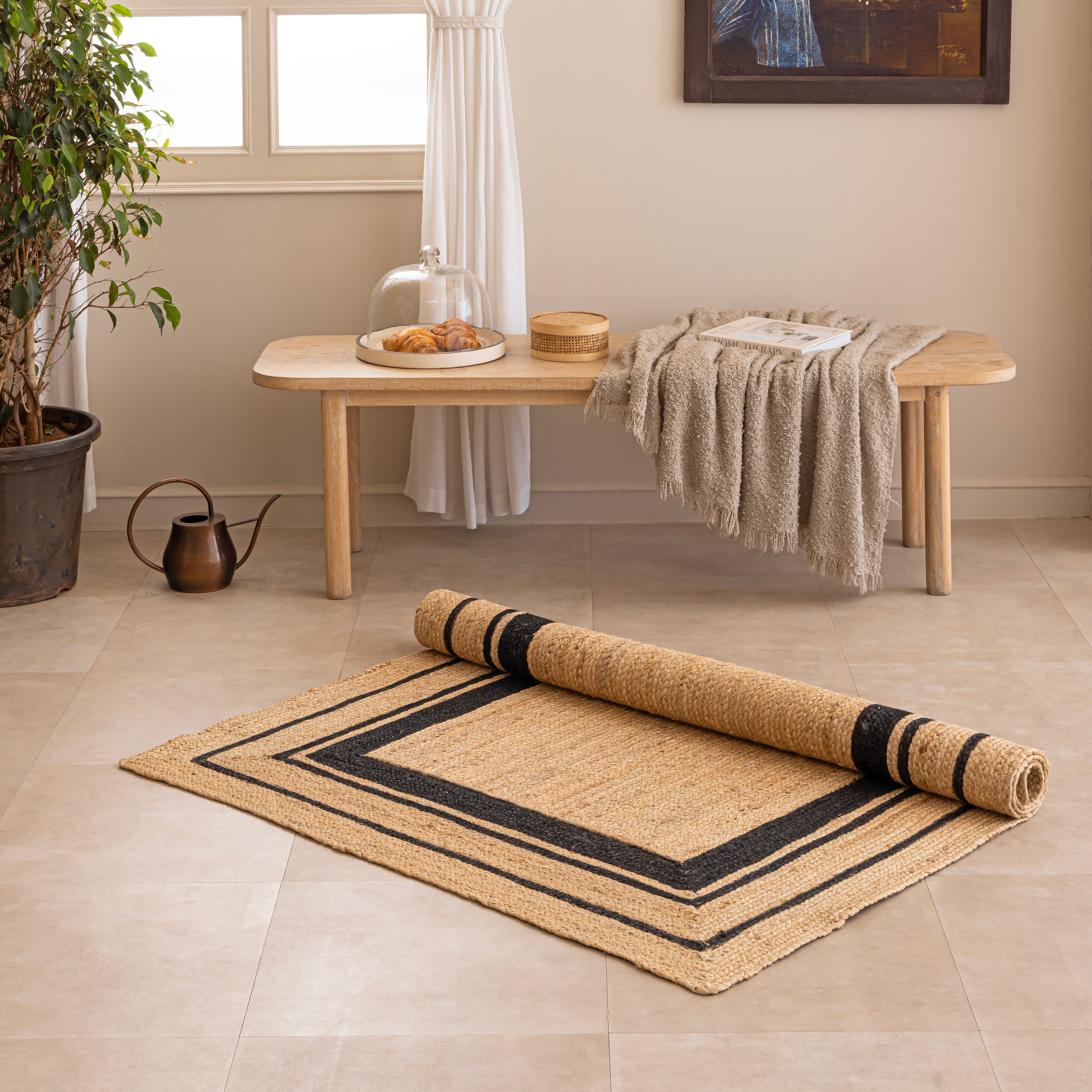 Handwoven natural tan jute area rug featuring a black double-striped border design, styled flat on a beige tiled floor next to a white ceramic vase with dried bunny tails and a hardcover book.