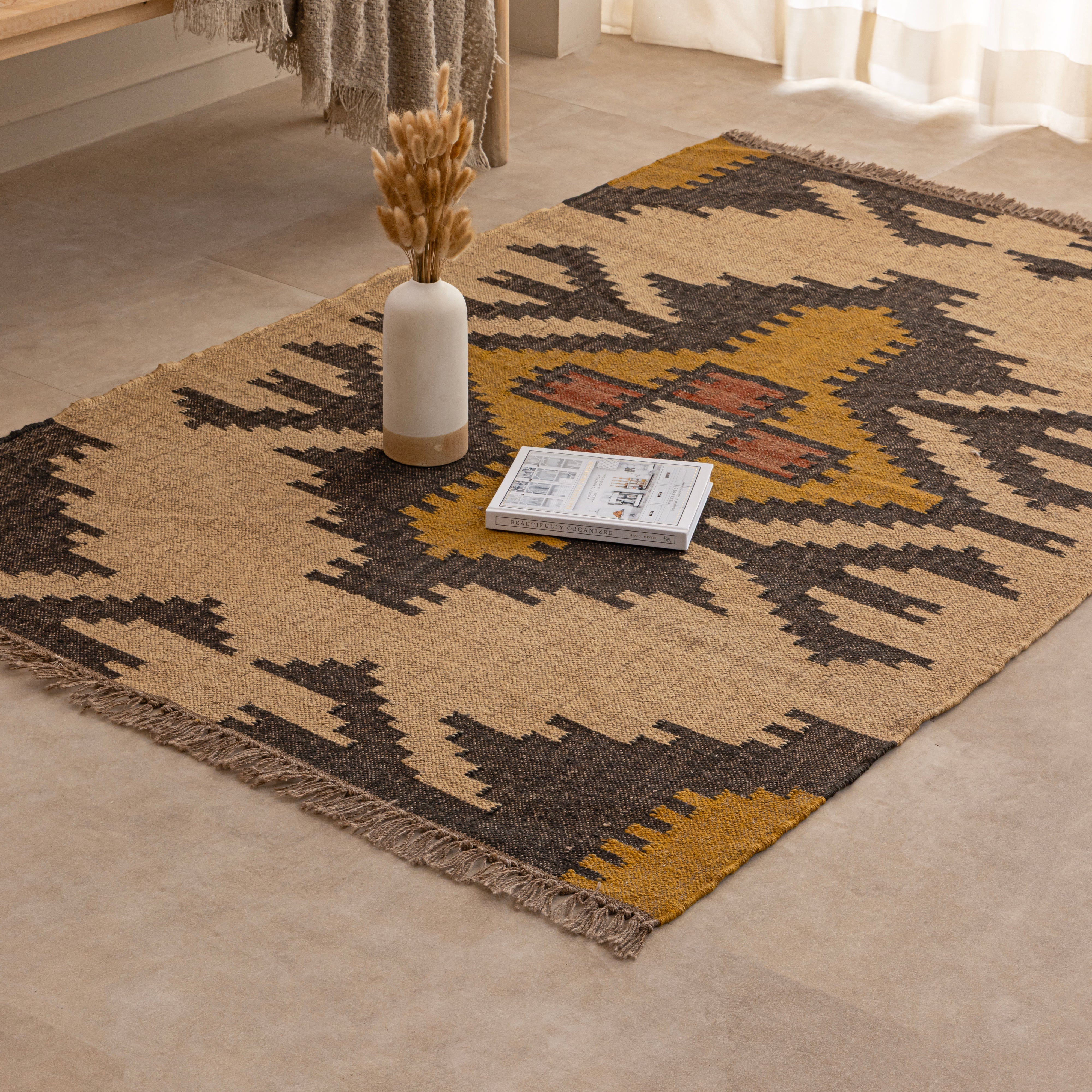 Handwoven Southwestern style natural jute kilim rug with geometric black and yellow patterns and fringed edges, styled on a beige tiled floor in front of a wooden bench and large indoor plant.