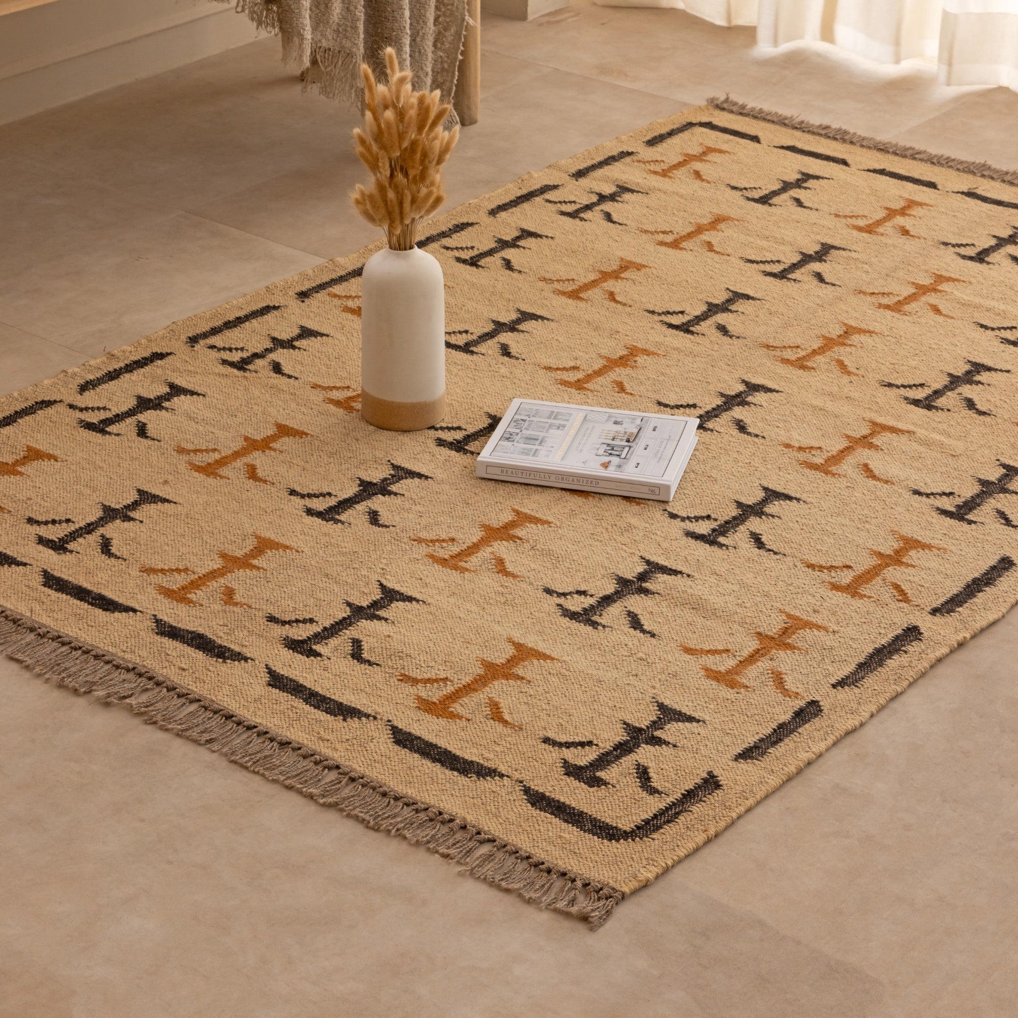 Handwoven natural jute kilim rug in beige with black and rust geometric tribal patterns, fringed edges, styled with a vase and book in a warm neutral living space.