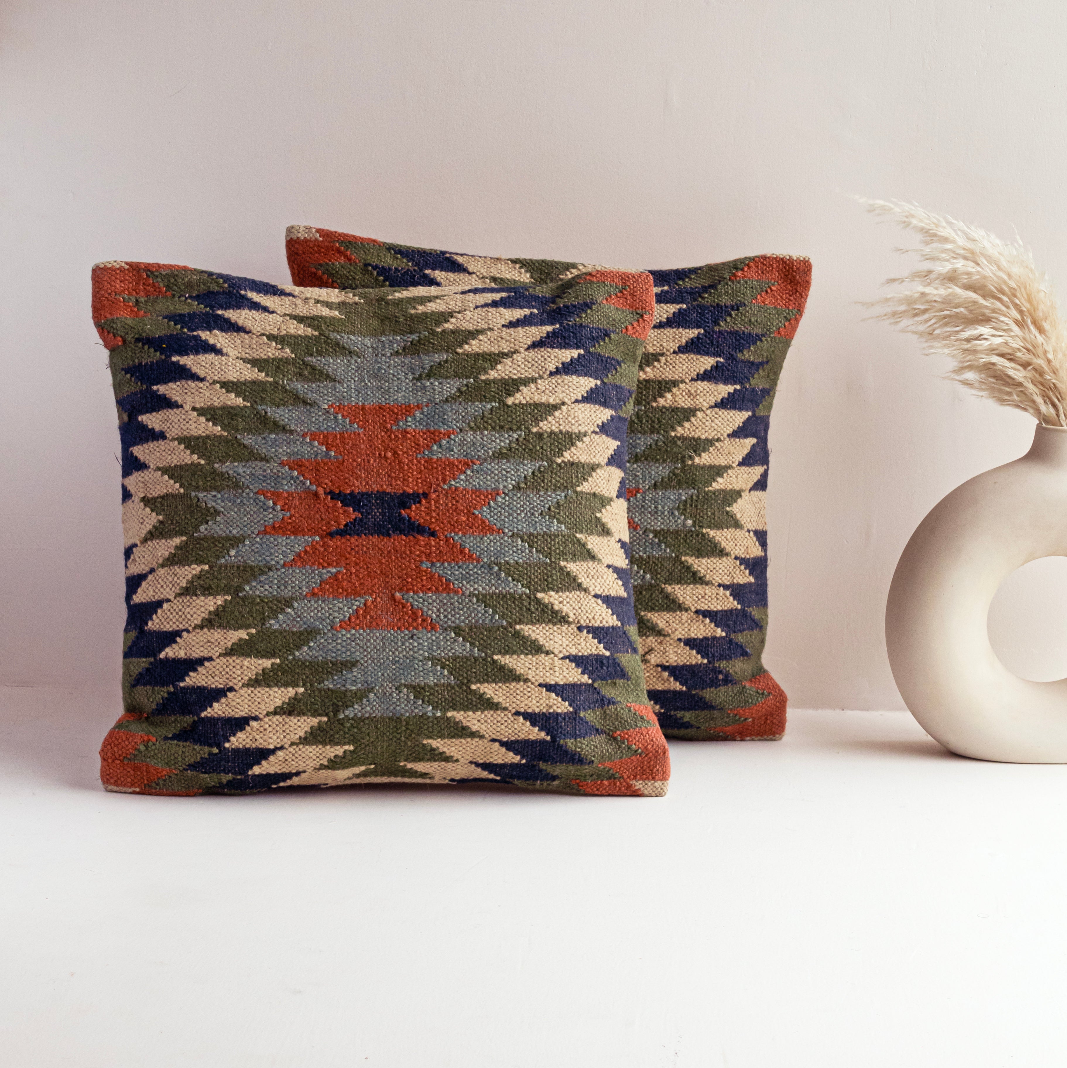 Handwoven Kilim cushion cover featuring a Southwestern tribal geometric pattern in rust, olive green, navy blue, and ivory displayed on a modern white tabletop.
