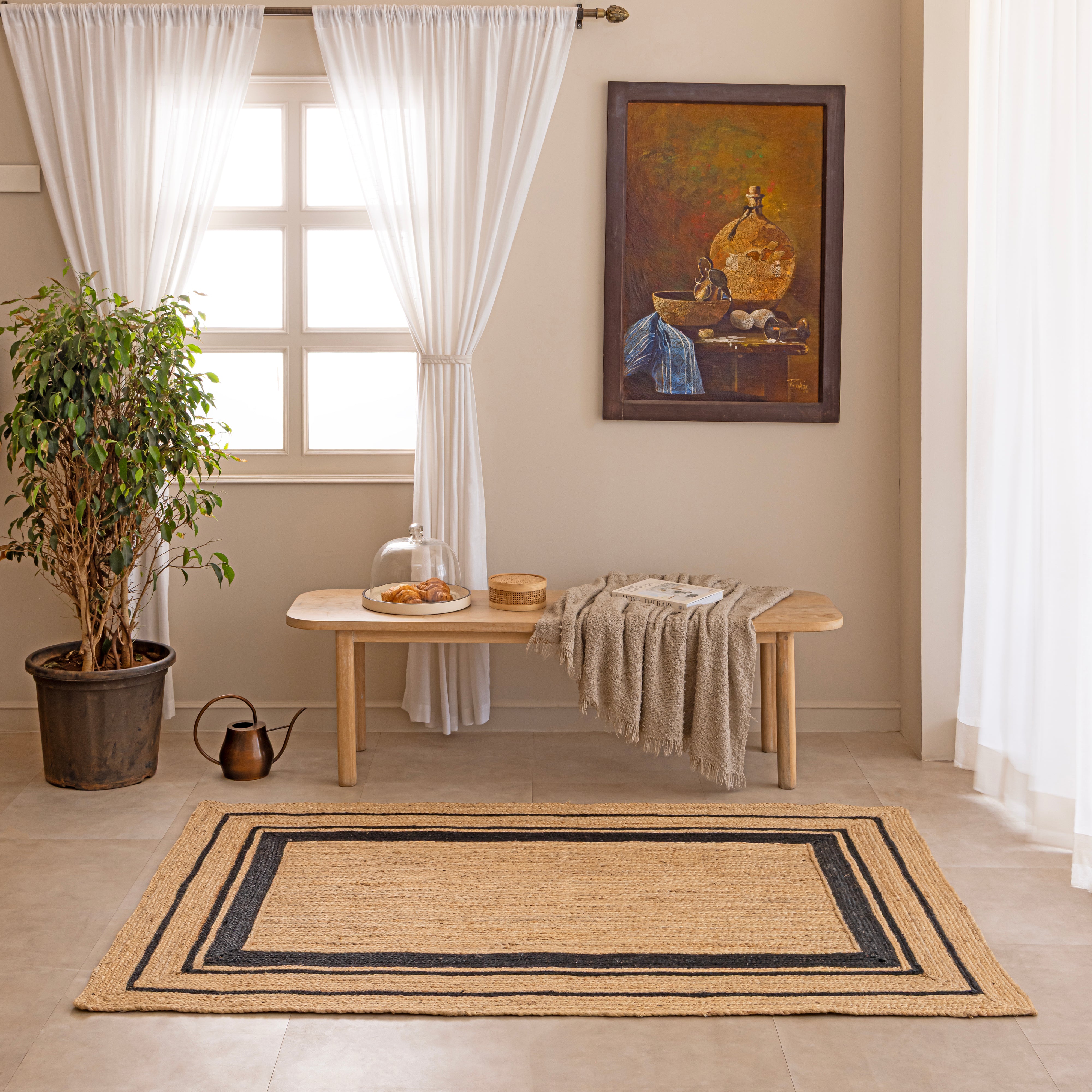 Handwoven natural tan jute area rug featuring a black double-striped border design, styled flat on a beige tiled floor next to a white ceramic vase with dried bunny tails and a hardcover book.