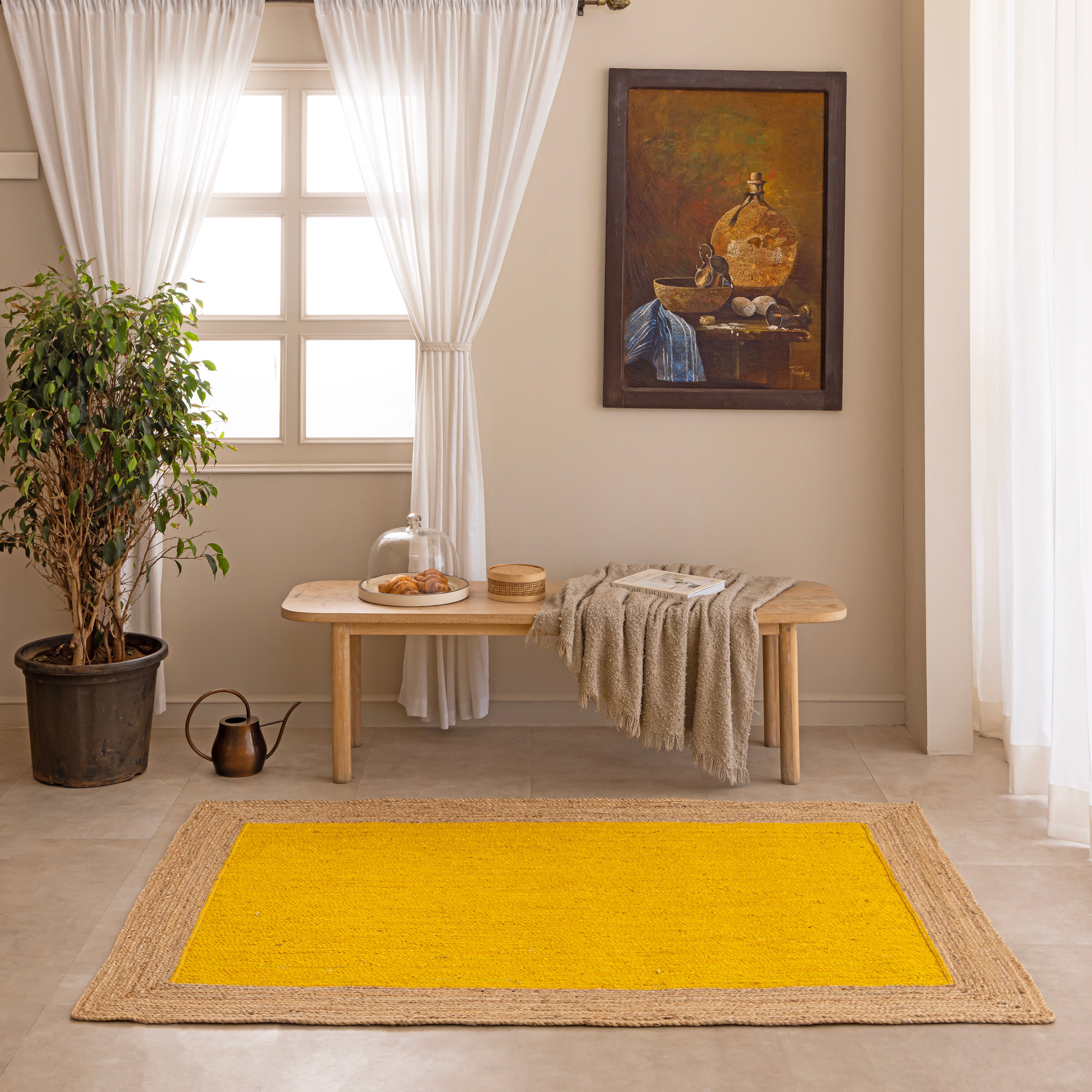Handwoven mustard yellow natural jute area rug with a braided tan border, laid flat on a light beige tiled floor alongside a ceramic vase with dried bunny tails and a book.