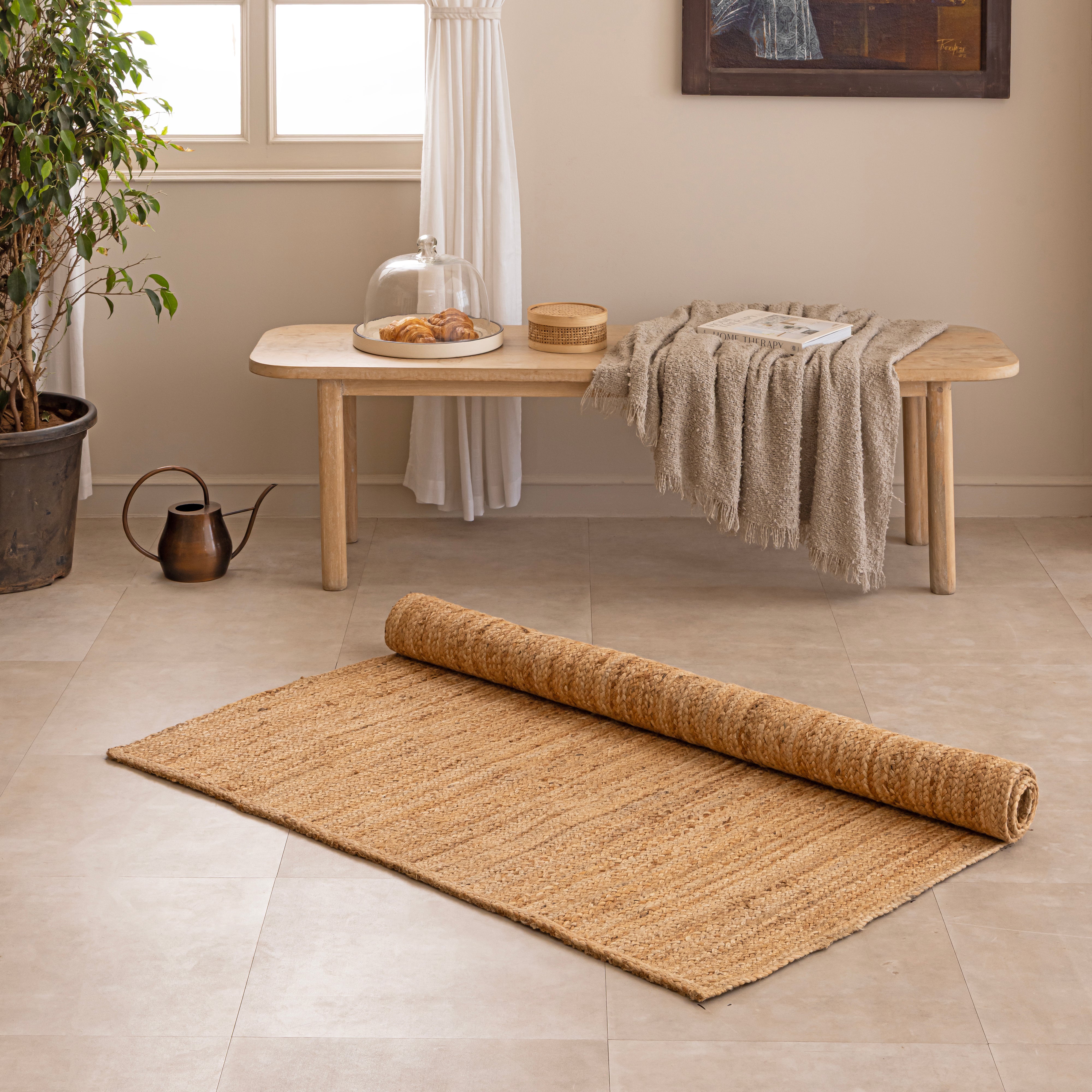 Handcrafted solid natural tan jute area rug featuring a thick braided texture, laid flat on a beige tiled floor beside a minimalist white ceramic vase with dried bunny tails and a hardcover book.