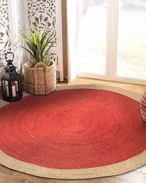 Handmade red round braided jute rug with a natural beige border, placed in a bright boho-style living room with indoor plants and wooden flooring.