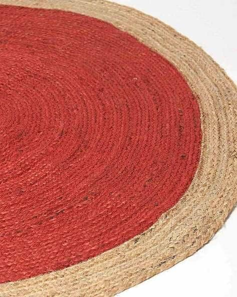 Handmade red round braided jute rug with a natural beige border, placed in a bright boho-style living room with indoor plants and wooden flooring.
